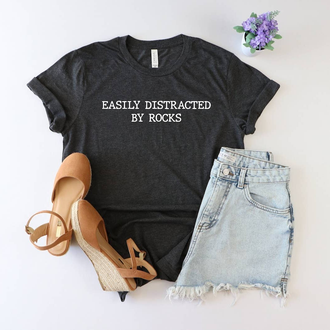 Black t-shirt with 'EASILY DISTRACTED BY ROCKS' text, sandals, and denim shorts on a light background