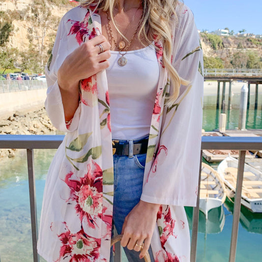 Woman wearing a floral kimono over a white top and blue jeans, standing by a waterfront with boats and mountains in the background.