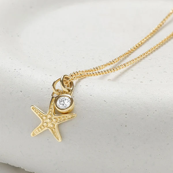 A gold starfish pendant necklace with a small clear round gemstone, displayed on a textured white surface.