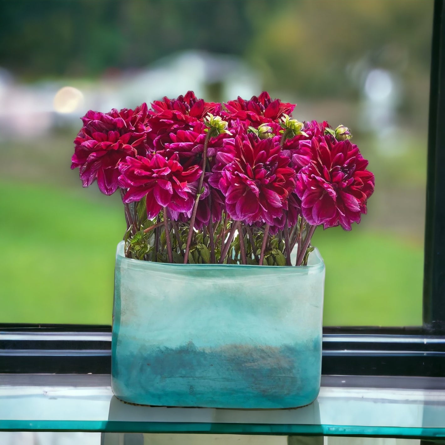 green and blue sea glass vase with dark pink flowers in it on a windowsill