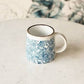 Mellow Monkey hand-stamped stoneware mug with blue floral design and gold rim detail. Microwave, dishwasher, and oven safe.