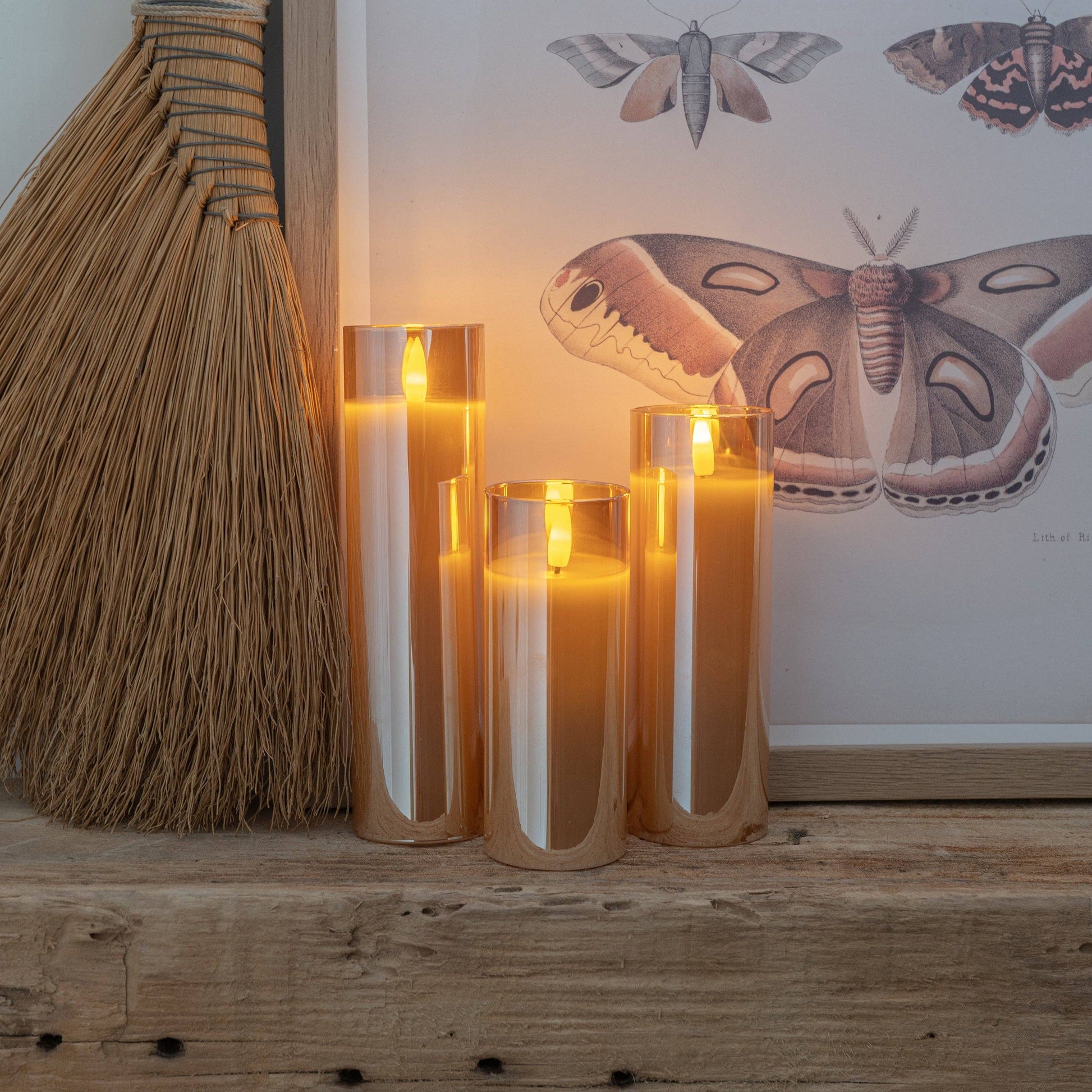 Mellow Monkey set of three amber glass flameless LED pillar candles with warm glowing light on a rustic wooden shelf.
