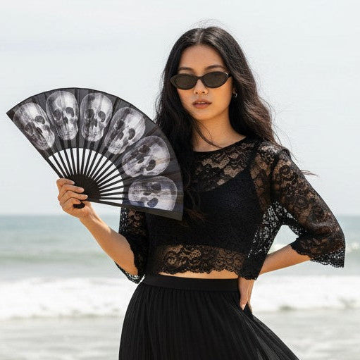 Woman in gothic black lace outfit holding a Mellow Monkey anatomical skull fabric fan on a sunny beach.