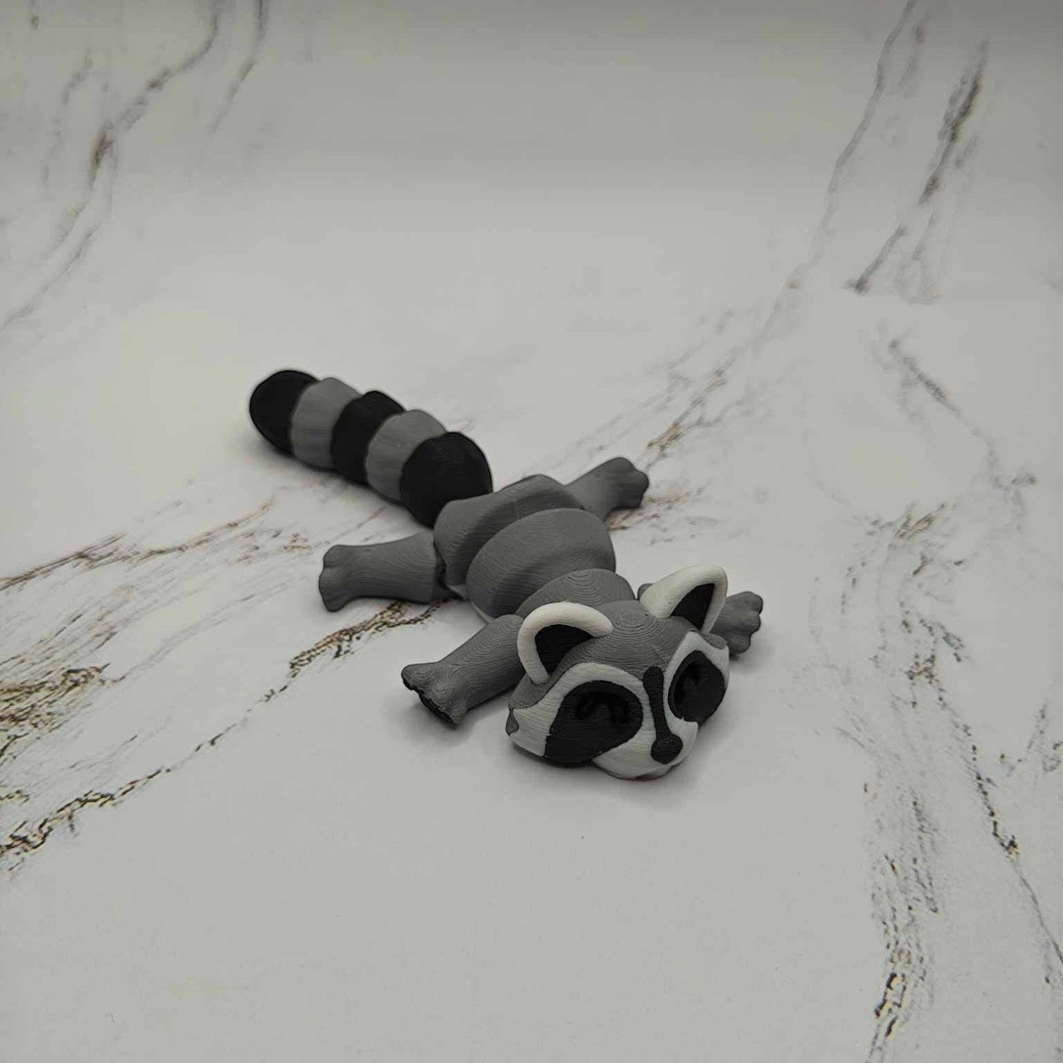 Mellow Monkey articulated grey and black 3D printed raccoon fidget toy lying flat on a white marble surface