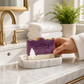 Mellow Monkey handmade purple and cream artisan soap bar with pine tree design on a marble soap dish in a bathroom.