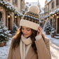 Mellow Monkey Aspen Fair Isle pom beanie in cream and blue worn by a woman in a snowy winter village street.