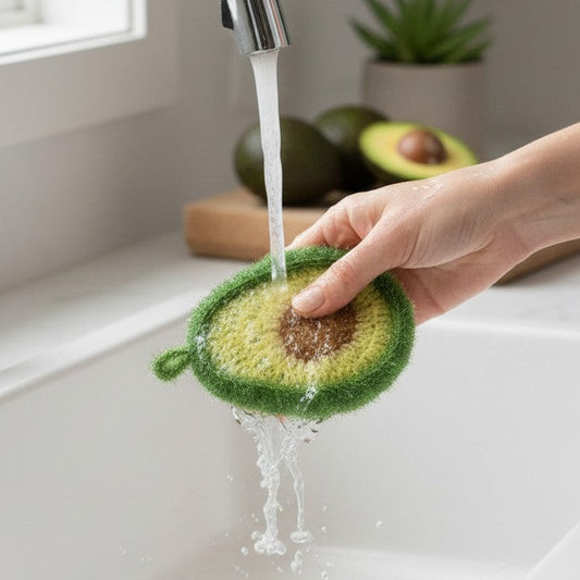 Mellow Monkey avocado-shaped dish scrubber being rinsed under a kitchen faucet in a white sink.
