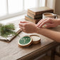 Mellow Monkey Wood Aromatics Balsam Pine hand salve in a round tin being applied to skin on a rustic wooden table.