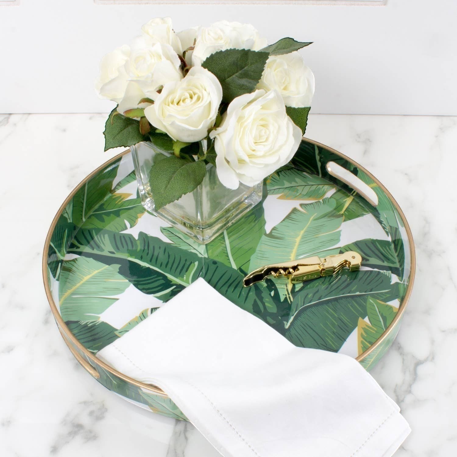 Mellow Monkey banana leaf print round resin serving tray with gold trim and handles, holding white napkins and a bouquet of white roses.
