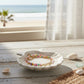 Mellow Monkey multi-color beaded anklet with gold chain resting in a white ceramic shell dish by the beach.