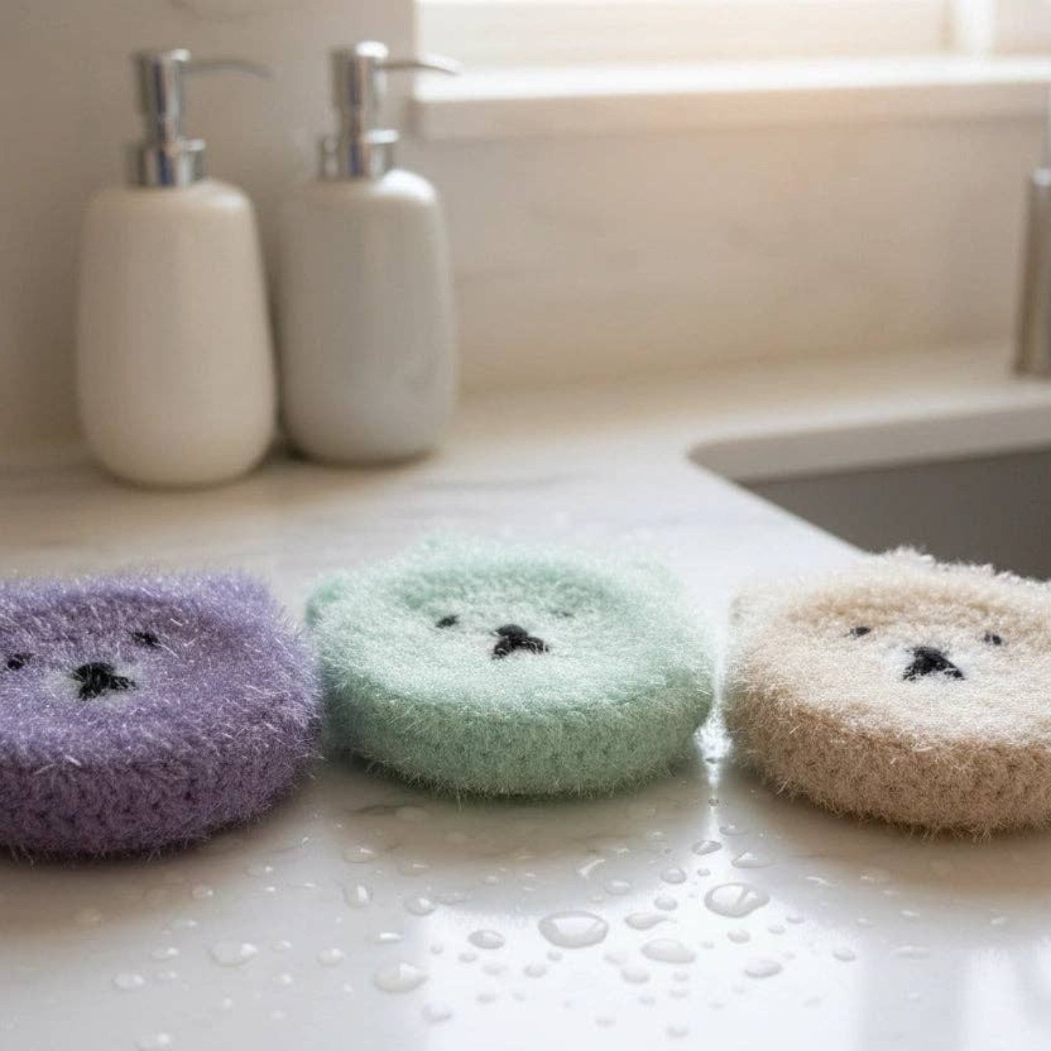 Mellow Monkey Good News Bears scrubby sponges in purple, aqua, and taupe. Cute bear face design with dual-sided scrubby and soft textures.