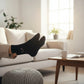 Mellow Monkey black crew socks with embroidered cat face featuring yellow eyes, worn while relaxing in a living room.