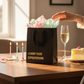 Mellow Monkey black kraft paper gift bag with gold foil text on a table with cake, candles, and a champagne glass.