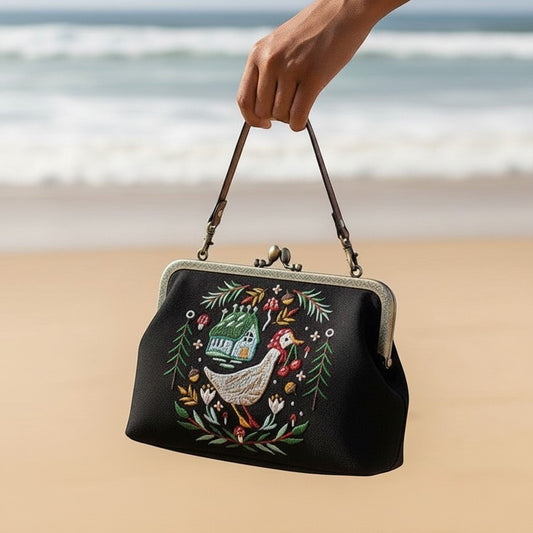 Mellow Monkey black linen kisslock bag with embroidered goose and cottage design held against a beach background