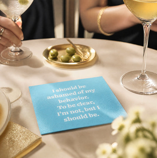 Mellow Monkey Carolina blue cocktail napkin with white text reading I should be ashamed of my behavior on a table.