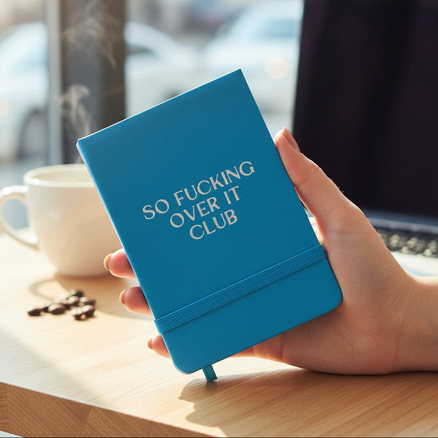 Mellow Monkey bright blue pocket notebook with So Fucking Over It Club text held in hand at a desk with coffee.