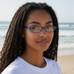 A woman wearing Mellow Monkey blue pocket reading glasses on a sunny beach with the ocean in the background.