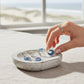 Mellow Monkey blue wave and seashell earrings in a rustic white dish held by a hand against a beach background