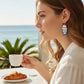 Mellow Monkey blue and white enamel fish earrings worn by a woman at a beachside cafe with coffee and a croissant.