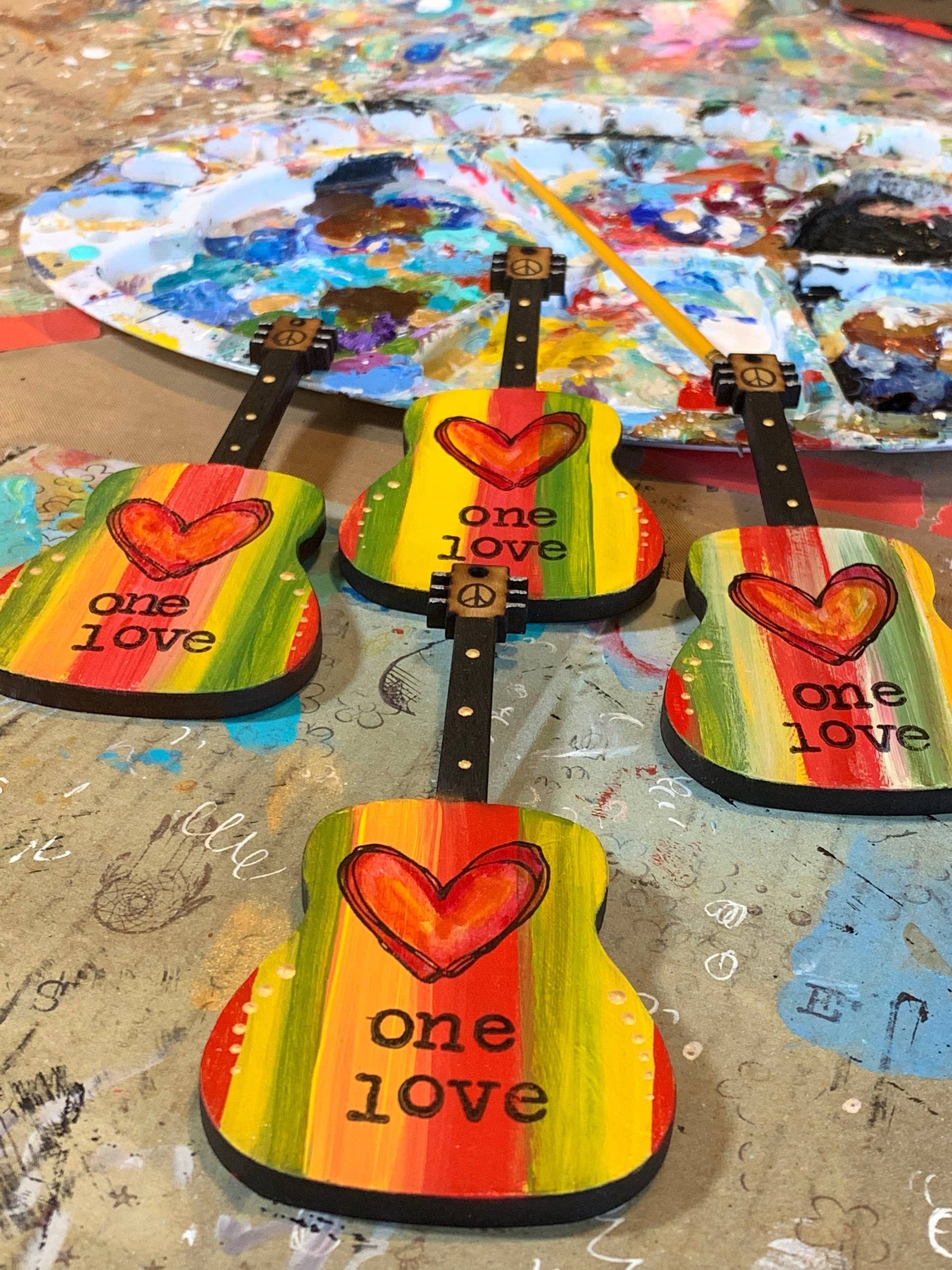 Mellow Monkey hand-painted wooden guitar ornaments featuring One Love text, red hearts, and Rasta-inspired stripes.