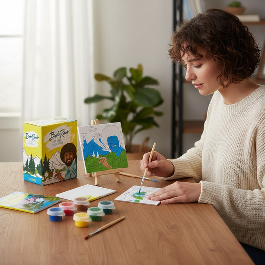 Mellow Monkey Bob Ross paint by numbers mini kit with canvases, paints, and easel being used by a woman at a table.