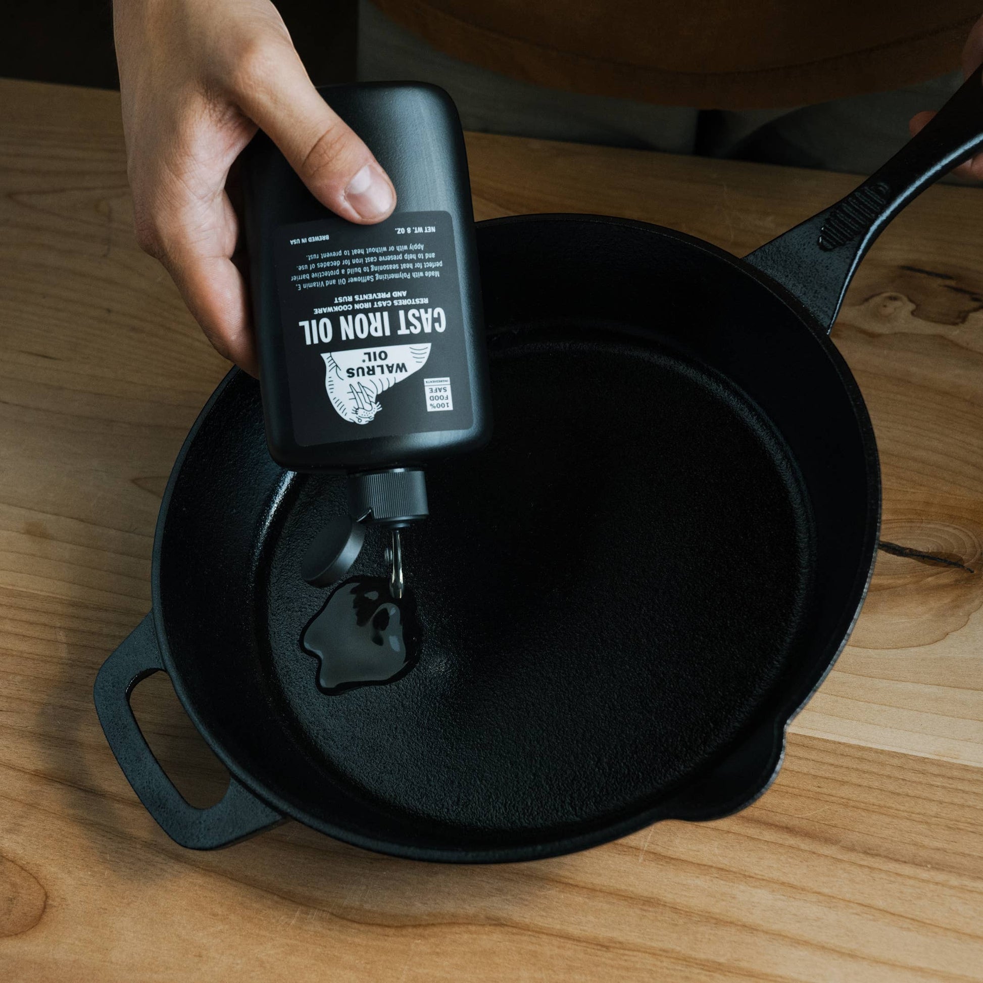 Mellow Monkey Cast Iron Oil 8-oz bottle being used to season a black cast iron skillet on a wooden surface.