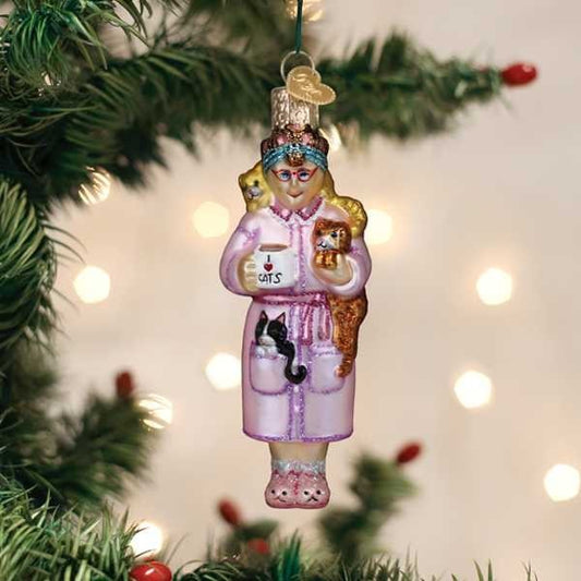 Mellow Monkey Cat Lady glass ornament with cats, holding a mug that says CATS. Pink robe, slippers, glasses.