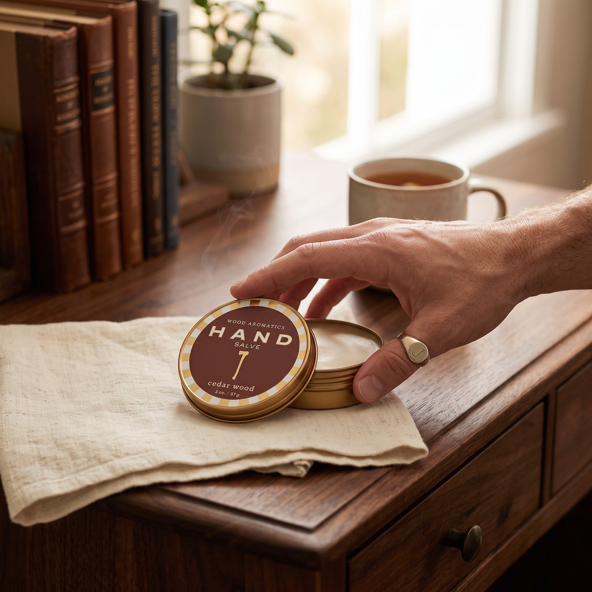 Mellow Monkey Wood Aromatics cedar wood hand salve in a round tin held by a hand on a wooden desk with books and tea.