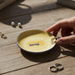 Mellow Monkey yellow ceramic trinket tray with a butter graphic and blue rim, held on a wooden table with jewelry.