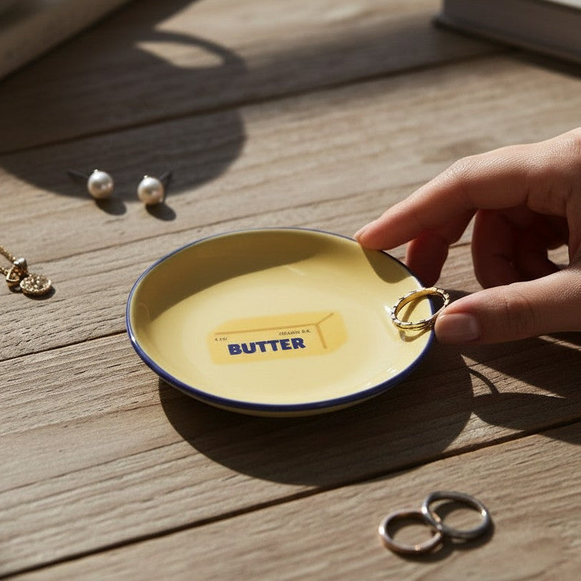 Mellow Monkey yellow ceramic trinket tray with a butter graphic and blue rim, held on a wooden table with jewelry.