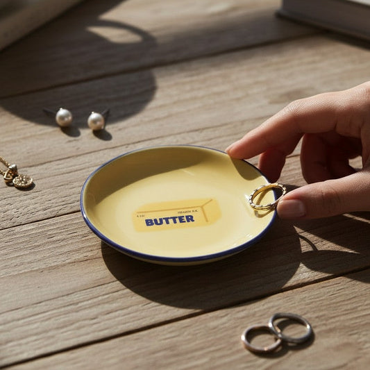 Mellow Monkey yellow ceramic trinket tray with a butter graphic and blue rim, held on a wooden table with jewelry.