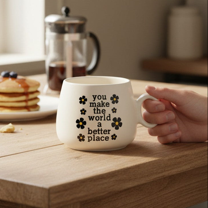 Mellow Monkey off-white ceramic mug with floral accents and the message You Make The World A Better Place on a table