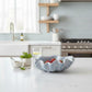 Mellow Monkey large ceramic shell bowl in dusty blue with reactive glaze, displayed on a kitchen counter with fruit.