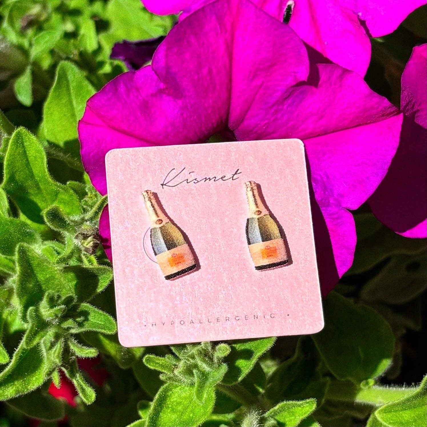 Mellow Monkey handmade champagne bottle stud earrings on a pink display card against a vibrant purple flower background.