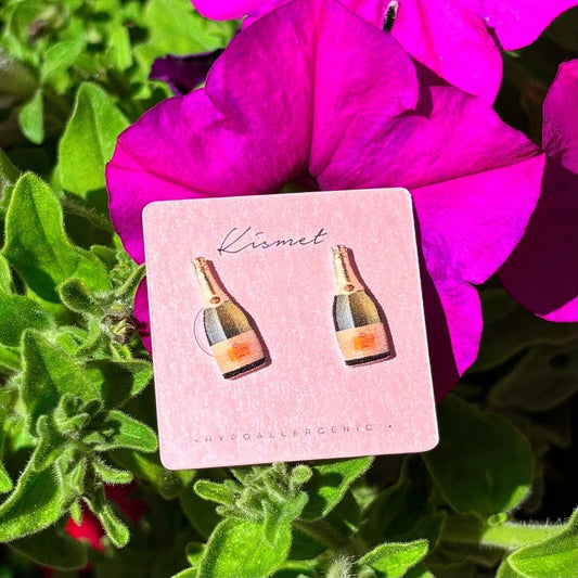 Mellow Monkey handmade champagne bottle stud earrings on a pink display card against a vibrant purple flower background.