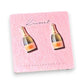 Mellow Monkey handmade champagne bottle stud earrings on a pink display card against a vibrant purple flower background.