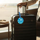 Mellow Monkey round blue luggage tag with white cloud pattern attached to a black suitcase in an airport lounge.