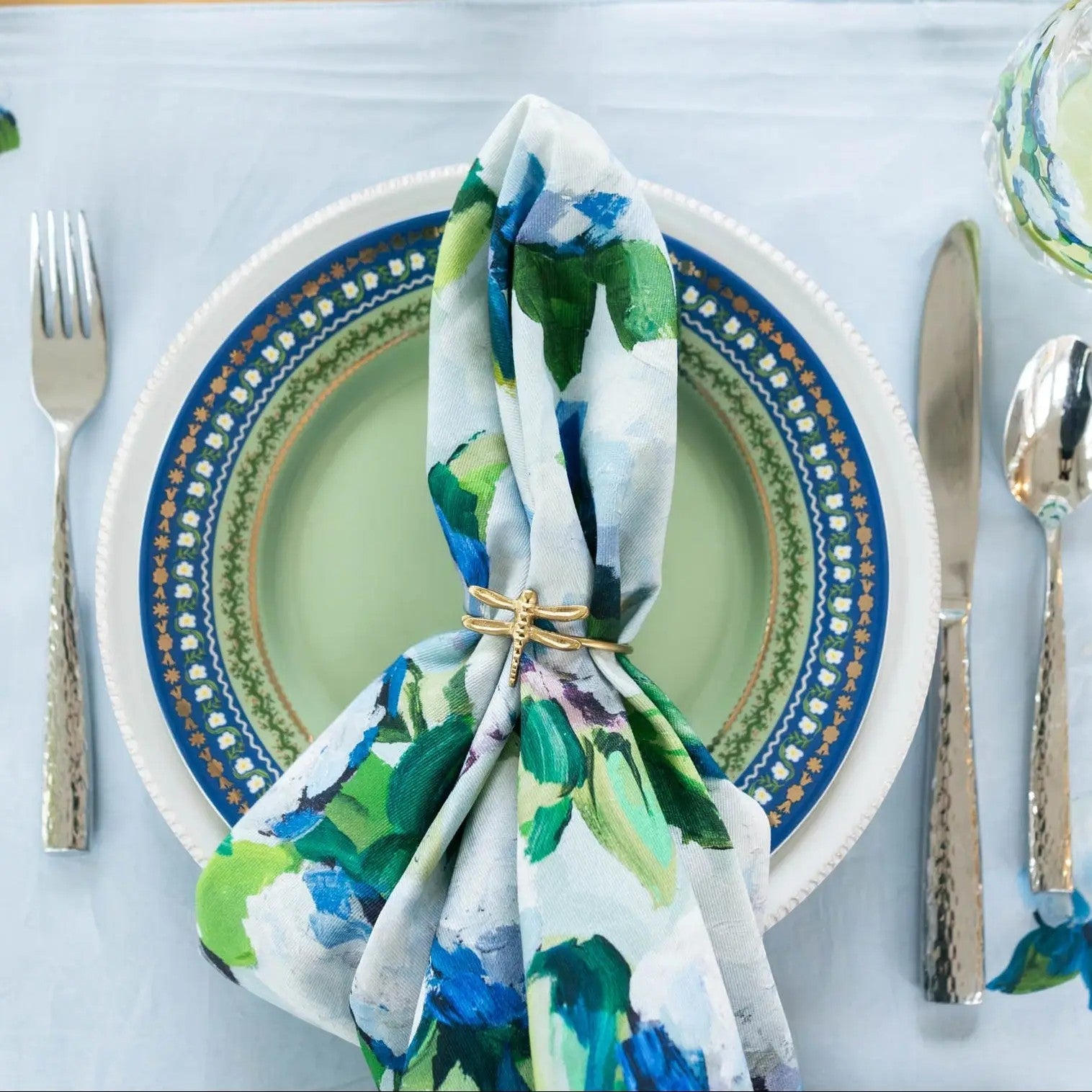 Mellow Monkey coastal hydrangea print cotton dinner napkin with a gold dragonfly napkin ring on a place setting.