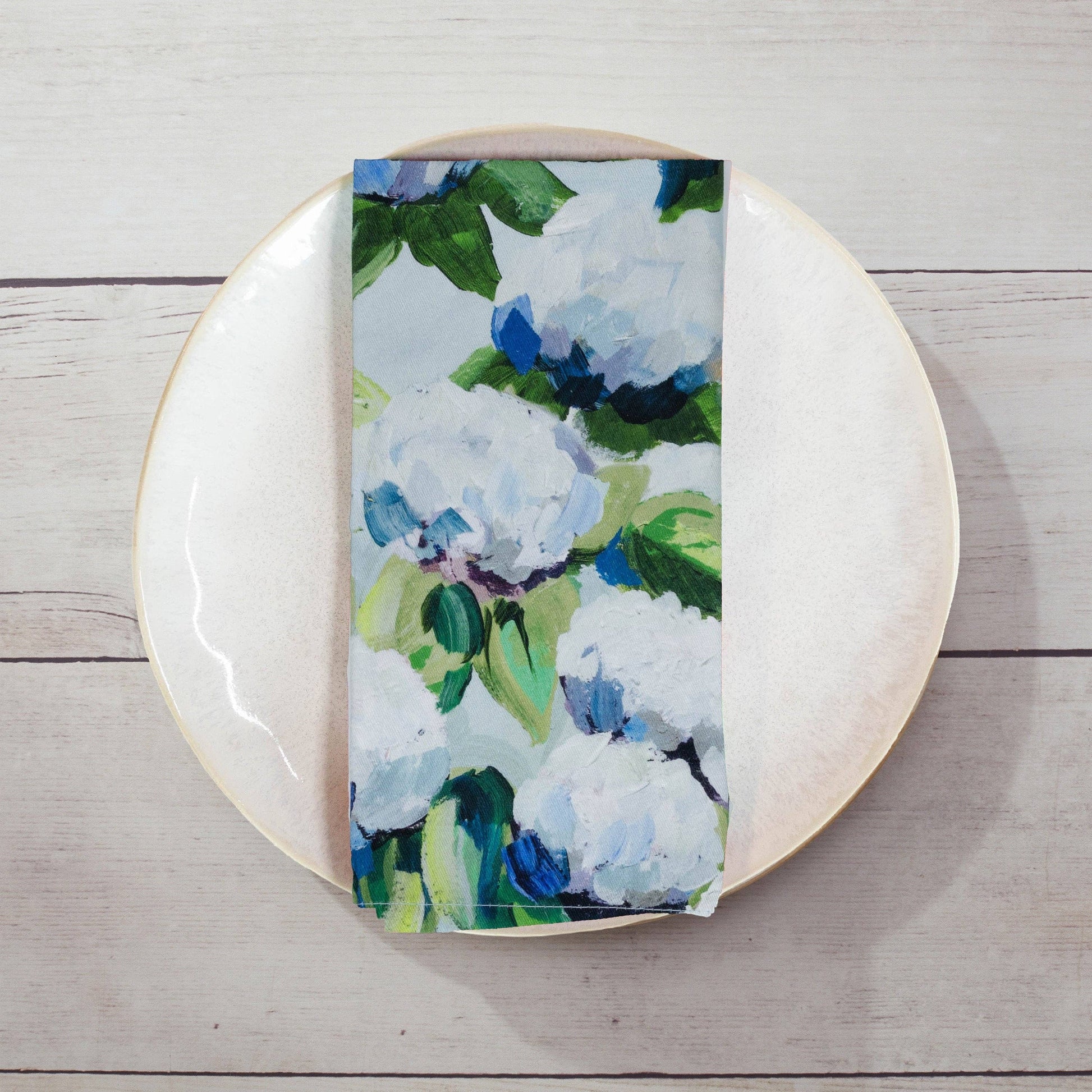 Mellow Monkey cotton dinner napkin featuring a coastal hydrangea floral print in blue, white, and green on a plate.