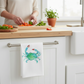 Mellow Monkey white cotton flour sack towel featuring a blue crab illustration hanging on a kitchen cabinet handle