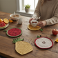 Mellow Monkey fruit shaped cotton crocheted pot holders on a wooden kitchen table with a person holding a tea cup