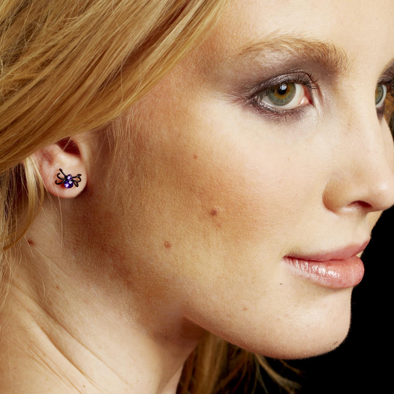 Mellow Monkey crystal spider stud earring worn on a woman's earlobe, featuring a small metal spider with a purple gem.