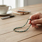 Mellow Monkey dark teal seed bead bracelet with 18K gold plated brass accents held in a hand on a wooden desk.