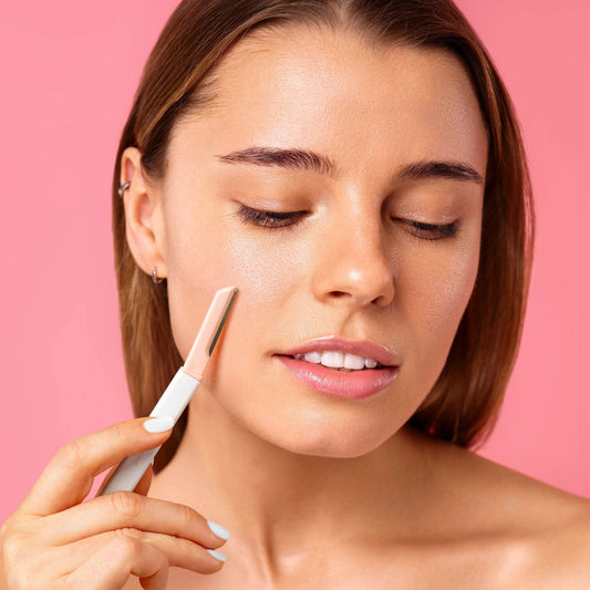 Mellow Monkey Fuzz Off Dermaplaner Facial Razor in pink and white, used by a woman on her cheek for smooth skin.