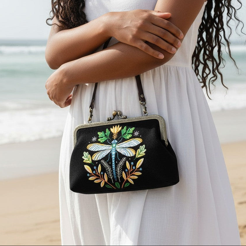 Mellow Monkey black cotton linen kisslock bag with embroidered dragonfly design held by woman on a beach