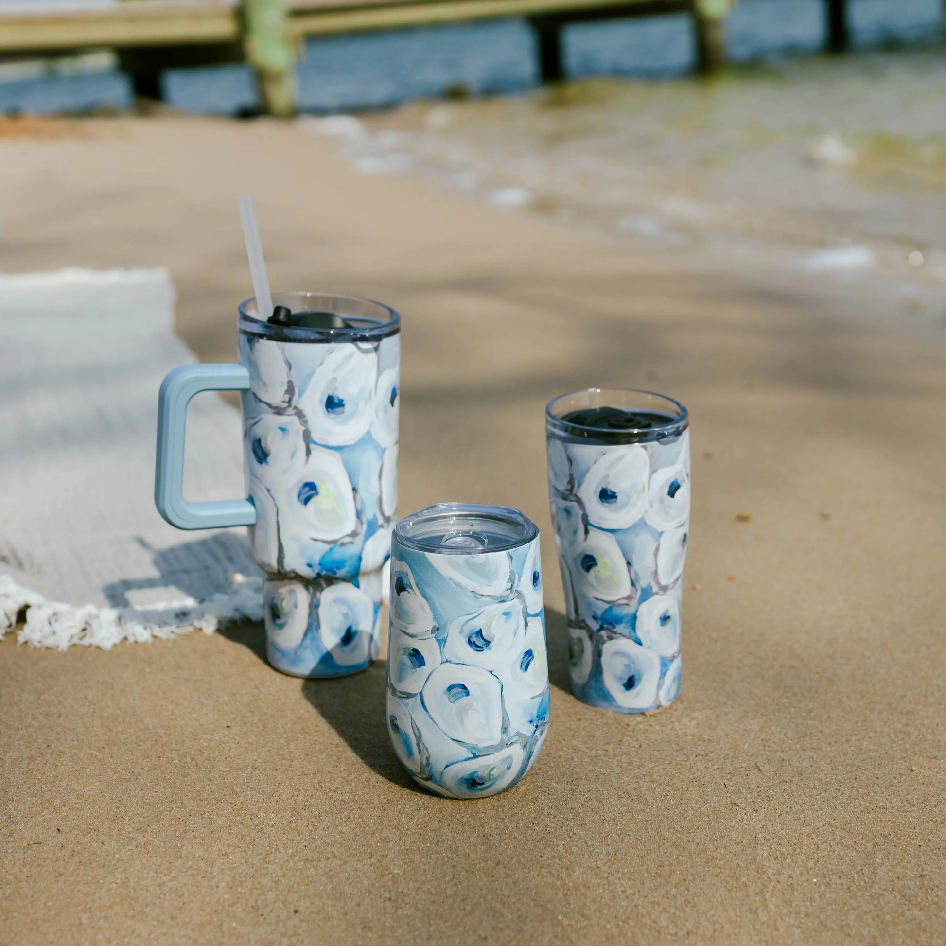 Mellow Monkey Ebb and Flow stainless steel wine tumblers with blue oyster shell pattern on a sandy beach.