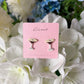 Mellow Monkey espresso martini stud earrings on a pink display card against a background of white flowers.