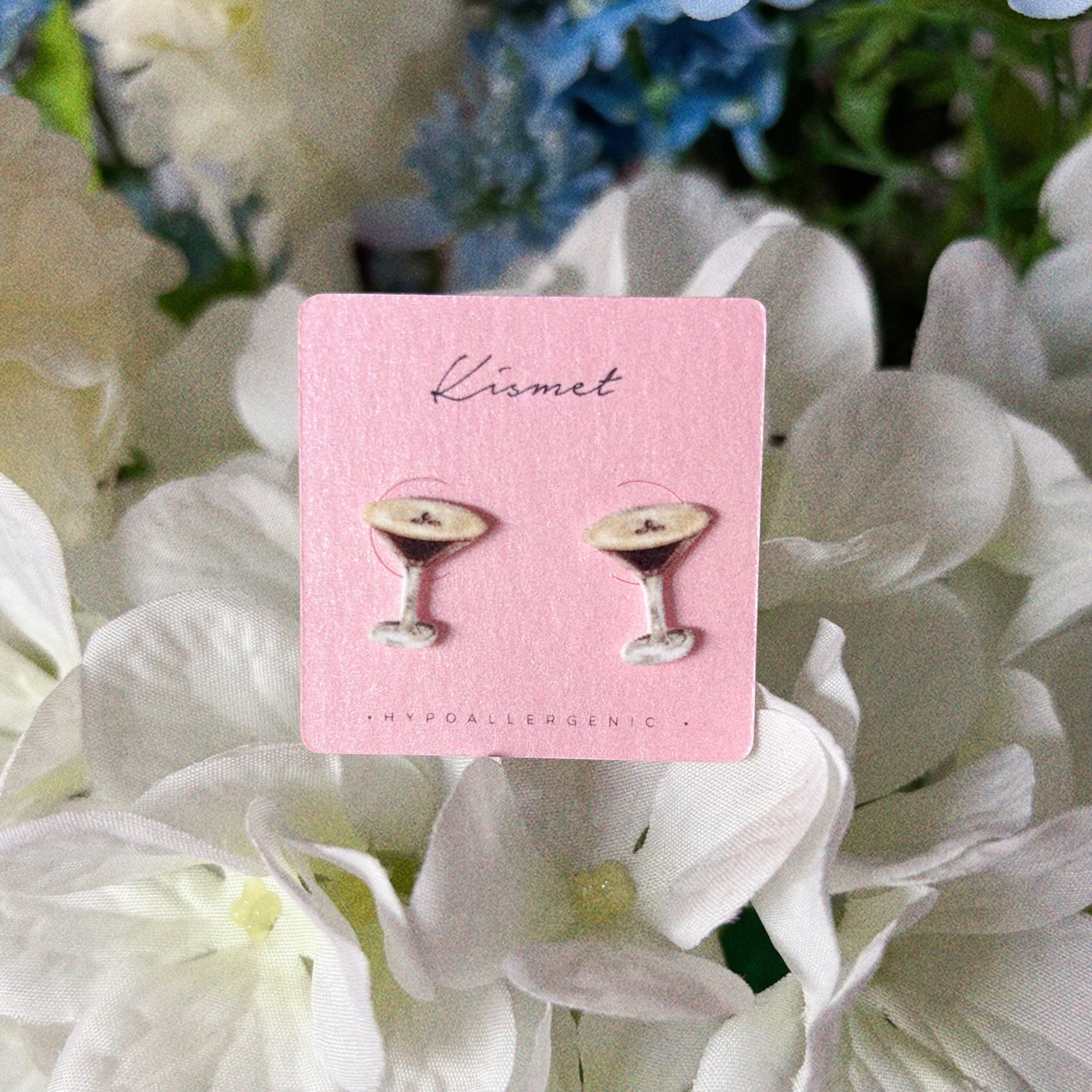 Mellow Monkey espresso martini stud earrings on a pink display card against a background of white flowers.