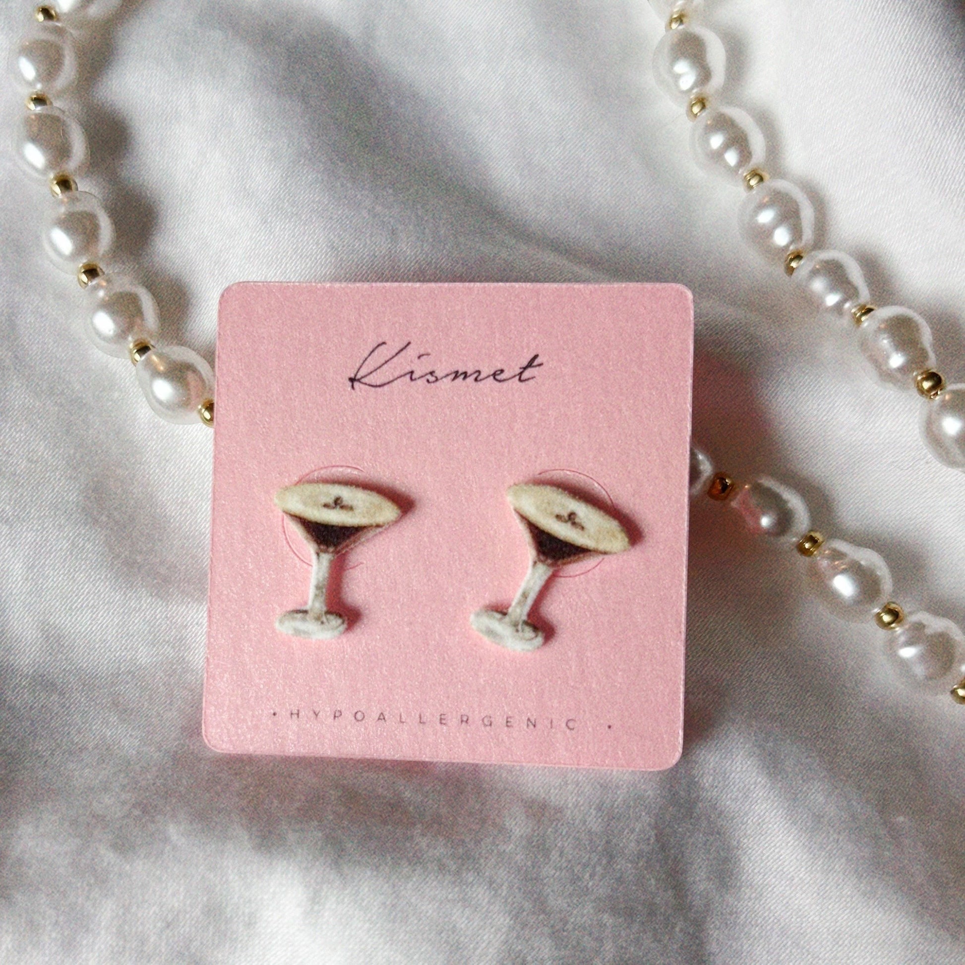 Mellow Monkey espresso martini stud earrings on a pink display card with a pearl necklace on white fabric.