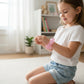 Young girl wearing a pink sparkly Mellow Monkey Feelzies sensory bracelet while sitting on a rug in a bright room.